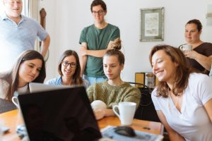 Seven people all gathered around a table, sitting and standing. Everyone is smiling except the person sat in the middle of the image, who looks sad and withdrawn